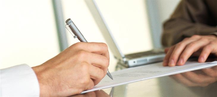 Eine Person mit weißem Ärmel schreibt mit einem Kugelschreiber etwas auf ein Blatt Papier. Eine andere Person sitzt gegenüber.