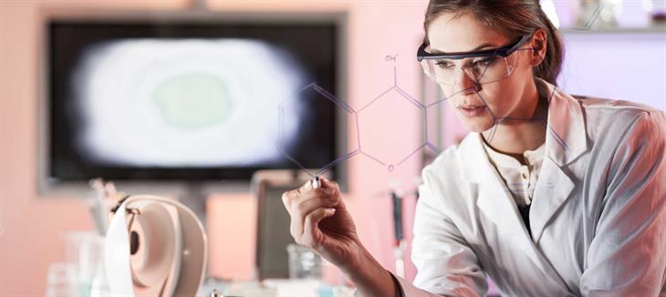 Eine junge Wissenschaftlerin in einem weißen Kittel mit einer Schutzbrille notiert eine chemische Strukturformel