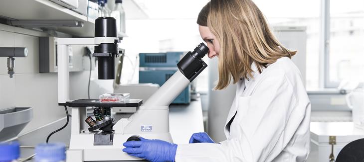 Eine Frau mit blonden Haaren und einem weißen Kittel sitzt an einem Mikroskop.