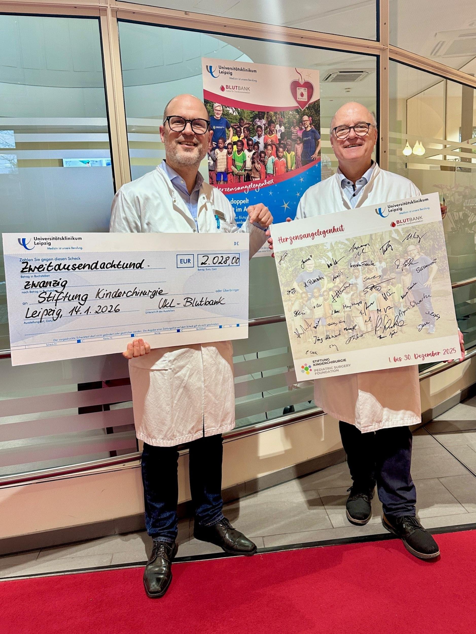 Prof. Martin Lacher (l.) freut sich über die Spendensumme der diesjährigen Aktion "Herzensangelegenheit" zugunsten der Stiftung Kinderchirurgie.  Prof. Reinhard Henschler präsentiert eine Tafel mit den Unterschriften von Blutspender:innen, die ihre Aufwandsentschädigung ganz oder anteilig gespendet haben.