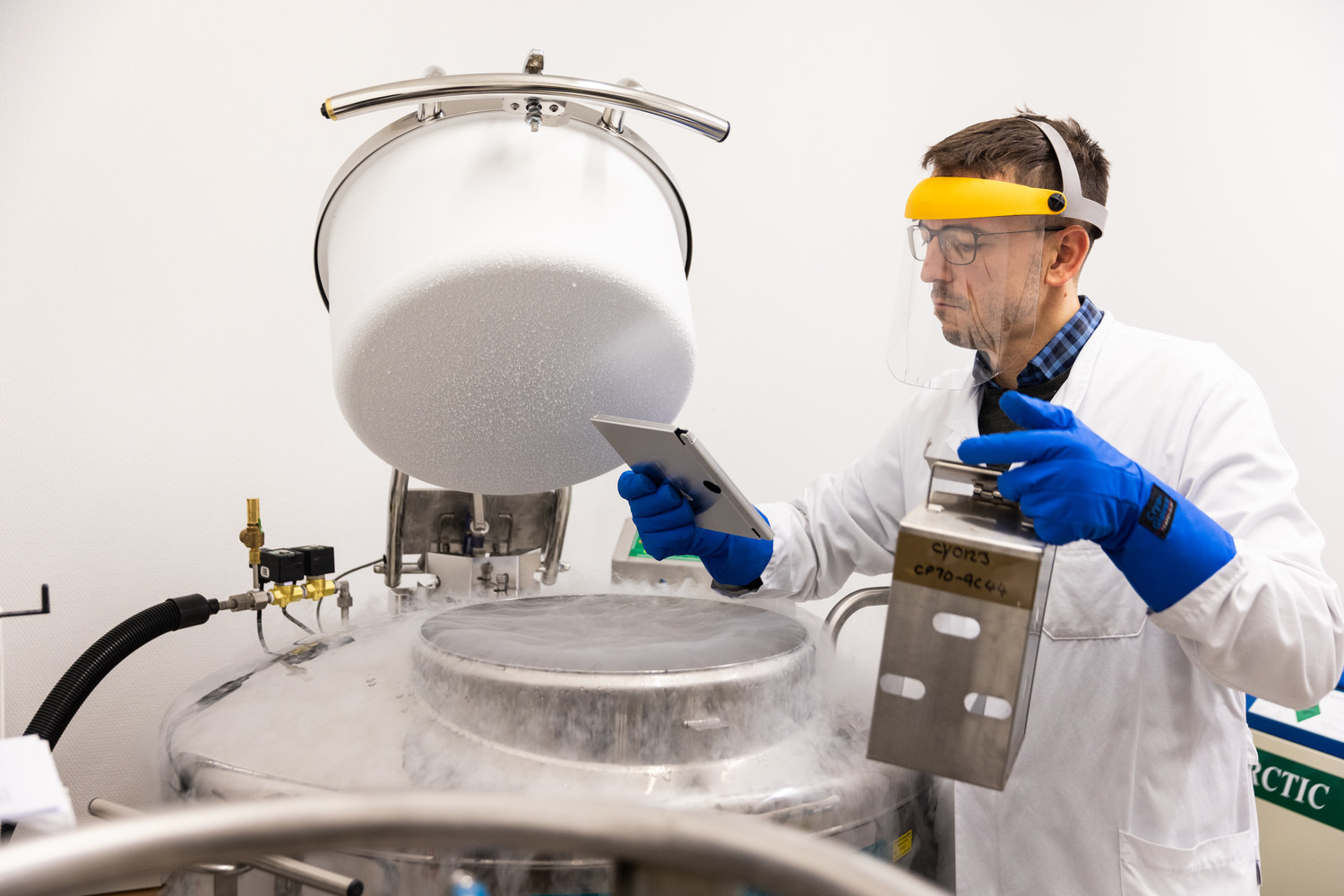 Dr. Vucinic in Schutzausrüstung vor einem sogenannten Kryotank und einer Kassette mit kryokonservierten, also tiefgefrorenen CAR-T-Zellen.