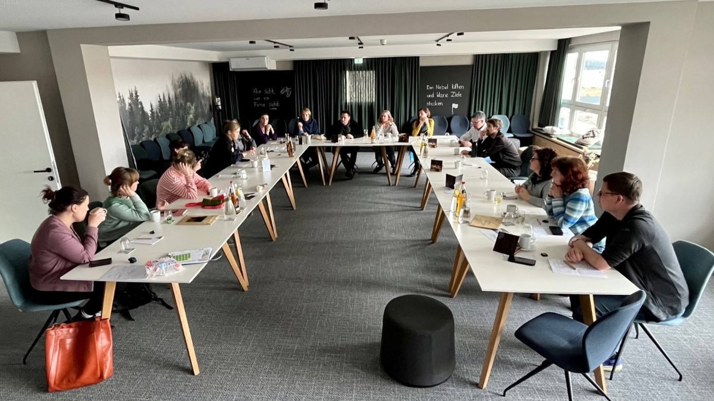 A group of people sit at long tables in front of a chalkboard that says " du hast alles und deine zeit "